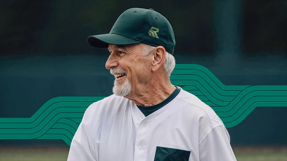 Older man with white hair and a beard wearing a green baseball cap and white baseball jersey smiling and looking to the side.