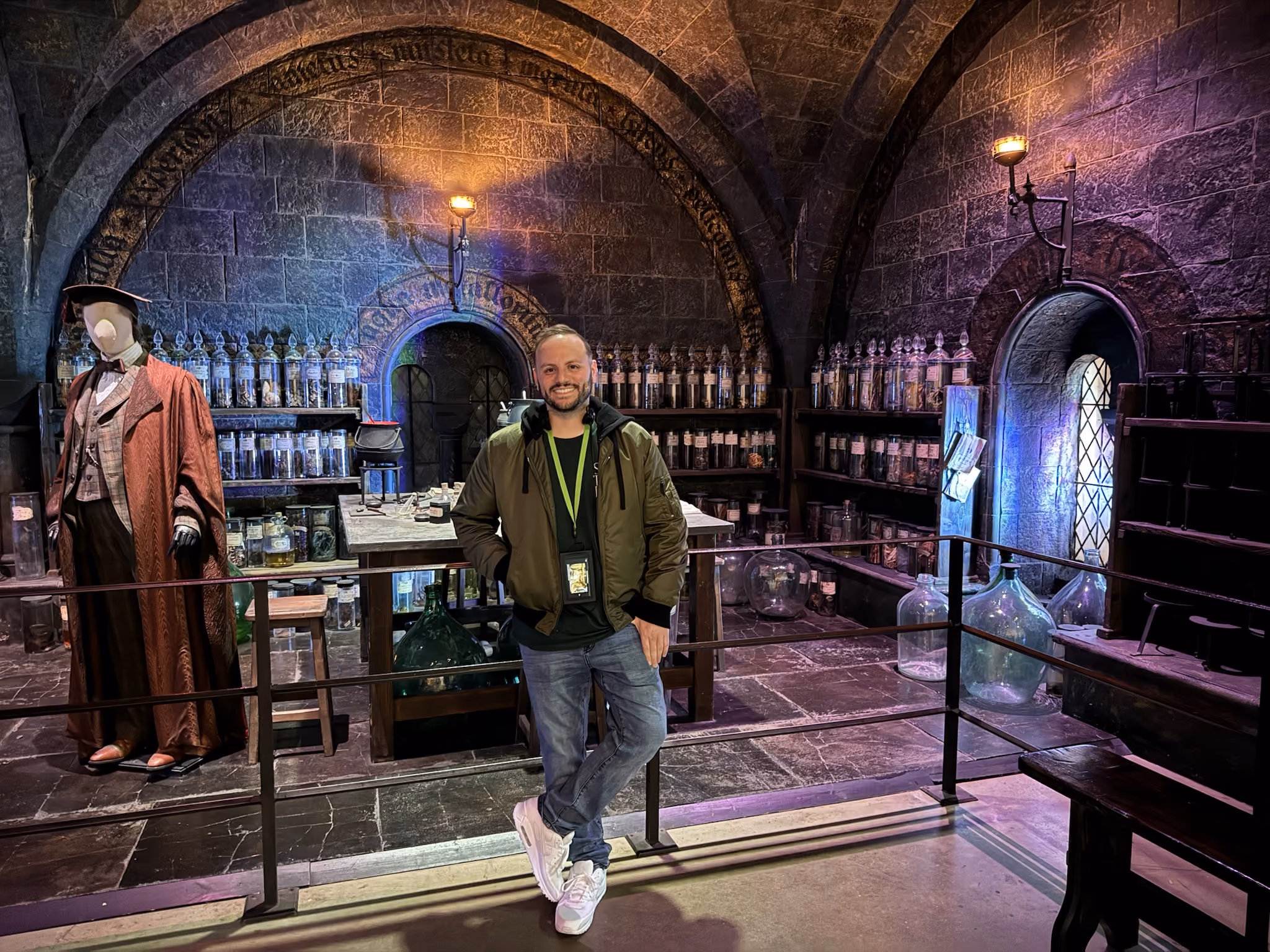 Smiling Greg wearing a green jacket and jeans leans against a metal railing in a dimly lit room resembling a medieval apothecary with stone walls, shelves filled with jars and bottles, and a mannequin dressed in period clothing.