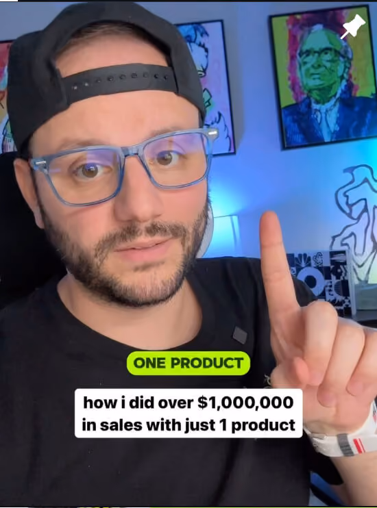 Greg with glasses and a backward black cap pointing upward with his index finger. Text overlay reads: 'ONE PRODUCT' and 'how I did over $1,000,000 in sales with just 1 product.' Colorful framed portraits and ambient lighting are visible in the background.