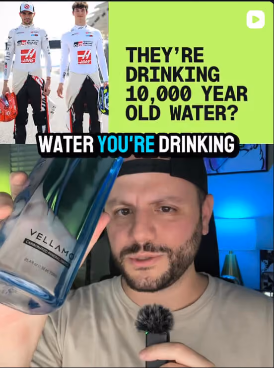 Greg wearing a black cap and beige shirt holds a Vellamo carbonated mineral water bottle and a microphone, with text above comparing drinking 10,000-year-old water by two Formula 1 drivers in racing suits and helmets.