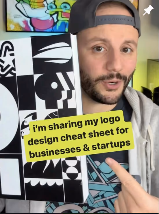 Greg wearing a backward black cap and gray hoodie holding a black and white logo design cheat sheet with abstract faces and shapes, pointing to text that reads 'i’m sharing my logo design cheat sheet for businesses & startups.'