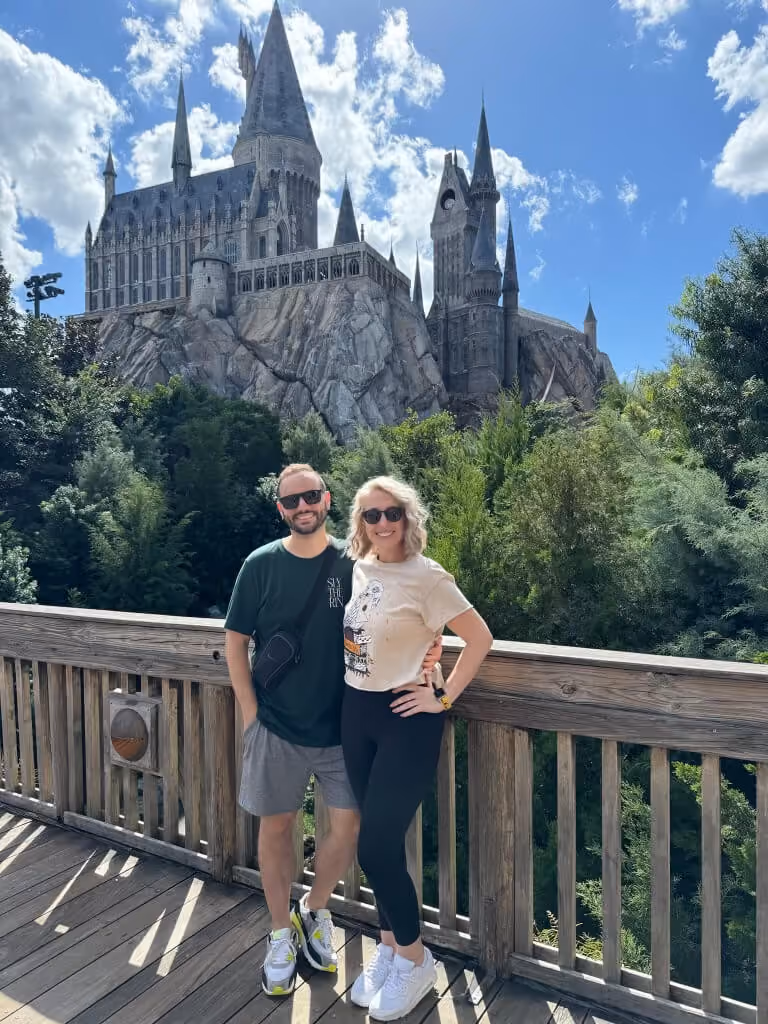 Greg and a woman wearing sunglasses pose smiling on a wooden bridge with green trees behind them and a large castle resembling Hogwarts in the background under a sunny sky with scattered clouds.