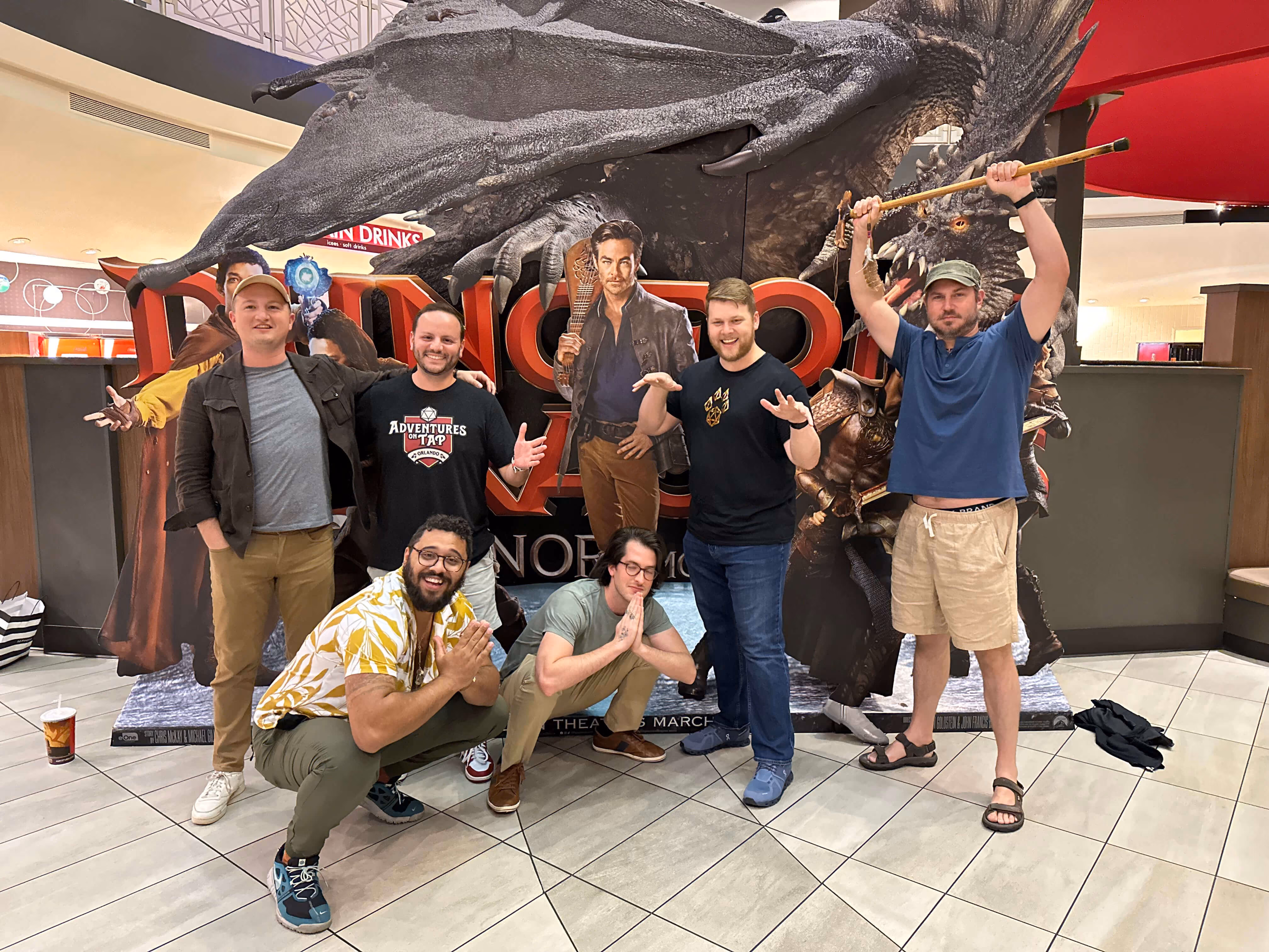 Six men posing at a mall in front of a large Dungeons & Dragons promotional display featuring a dragon and characters; two men are crouching, four standing, one holding a staff above his head, all smiling or making playful poses.