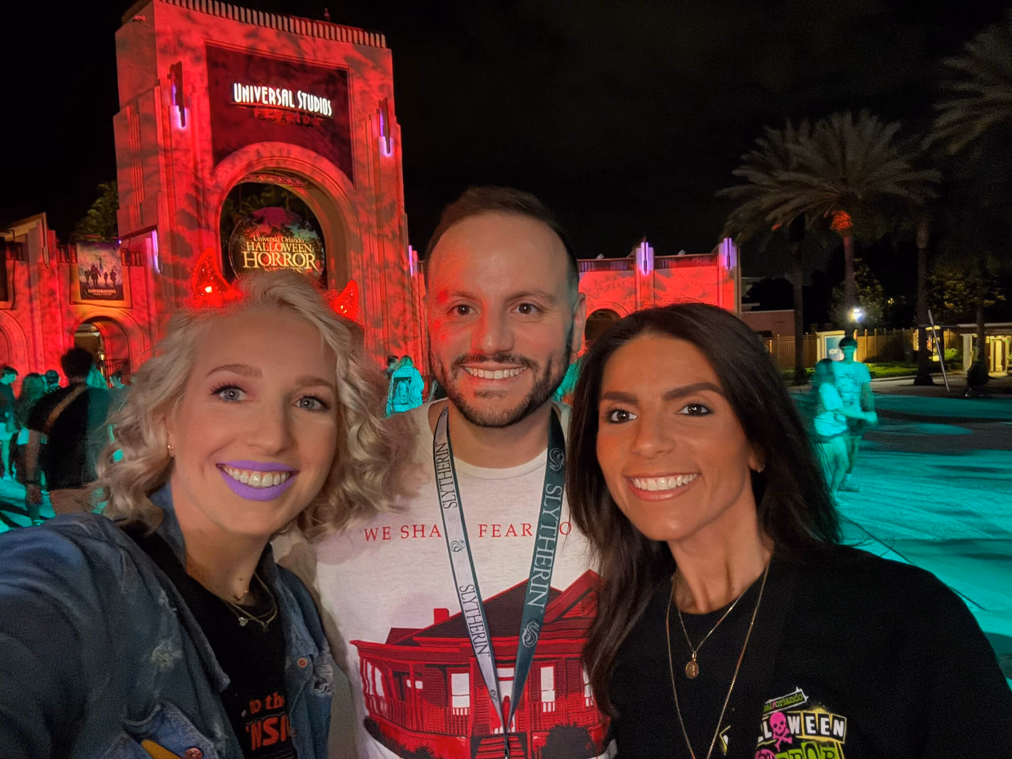 Three people smiling for a selfie at Universal Studios Halloween Horror event at night; the background building is lit in red with 'Universal Studios' and 'Halloween Horror' signs.