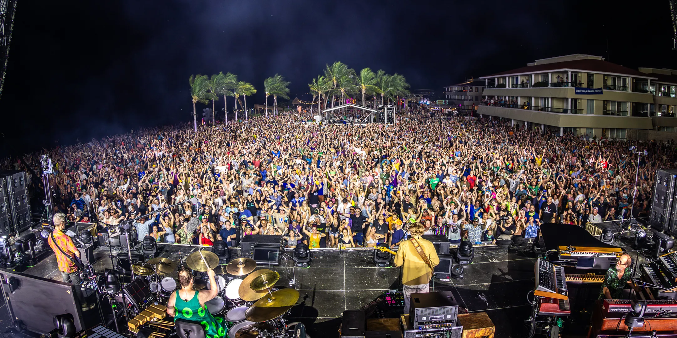 A lively concert scene with a large, enthusiastic crowd facing the stage under a night sky. Palm trees