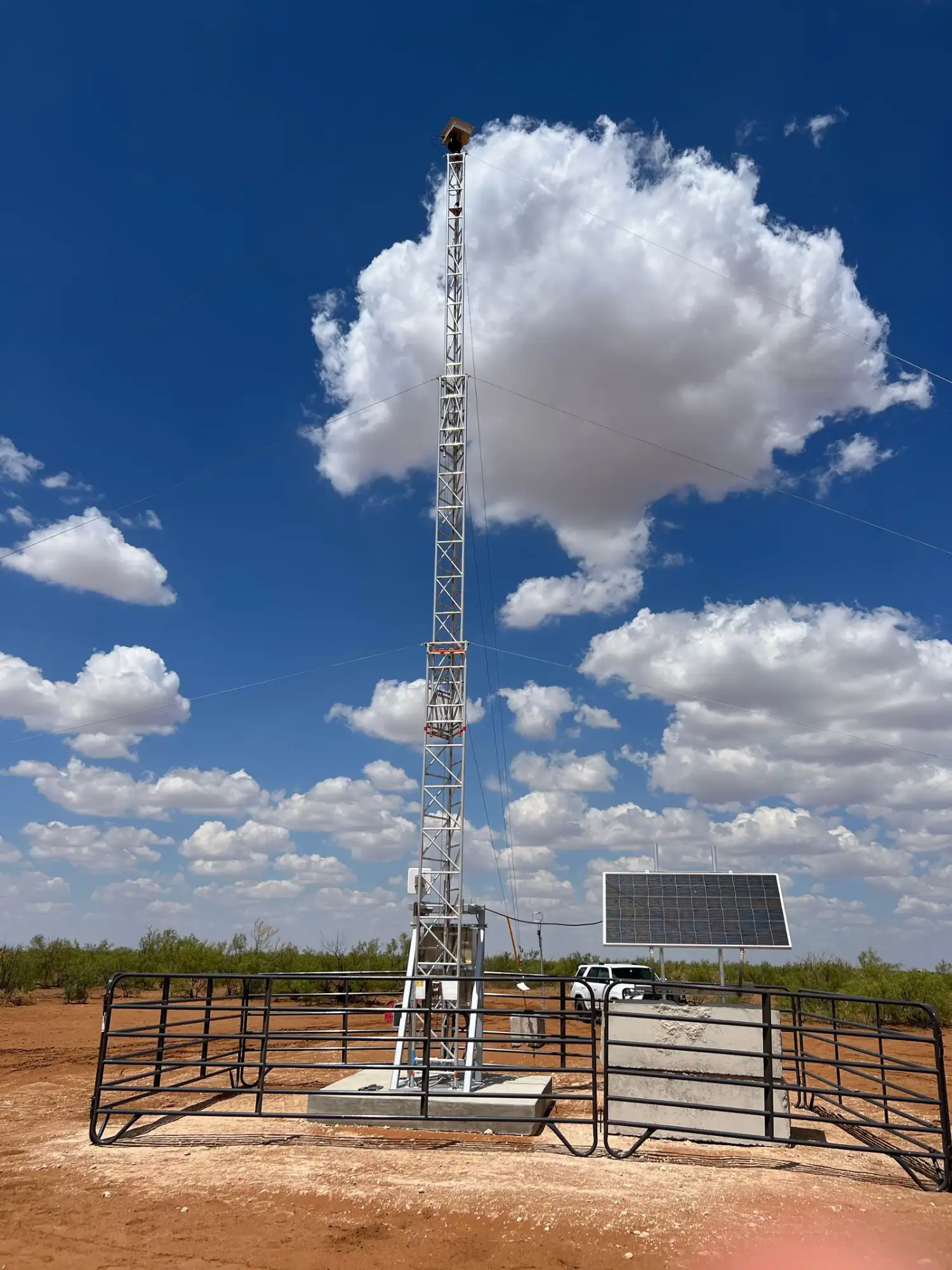 Tall metal weather monitoring tower with multiple instruments, surrounded by a black metal fence, with solar panels and a white vehicle in the background under a blue sky with clouds.