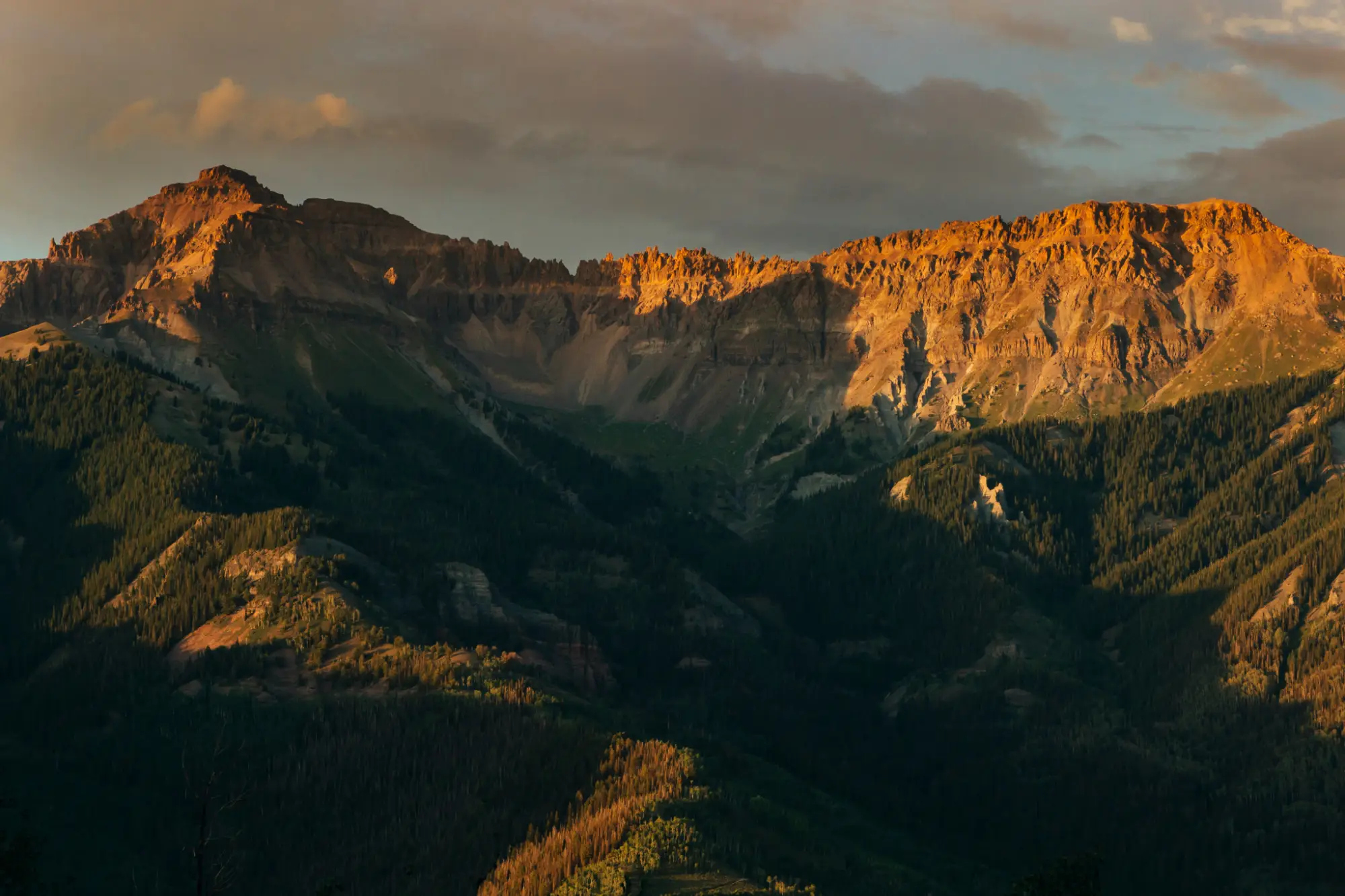 Mountain range bathed in golden sunlight with forested lower slopes under a cloudy sky.