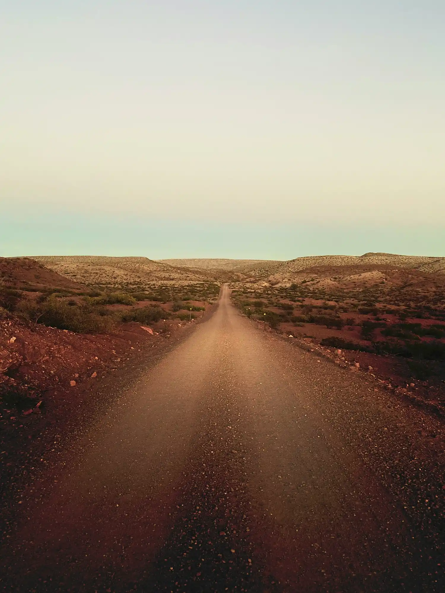 Long dirt road stretching through a dry, rocky desert landscape with low hills under a clear sky.