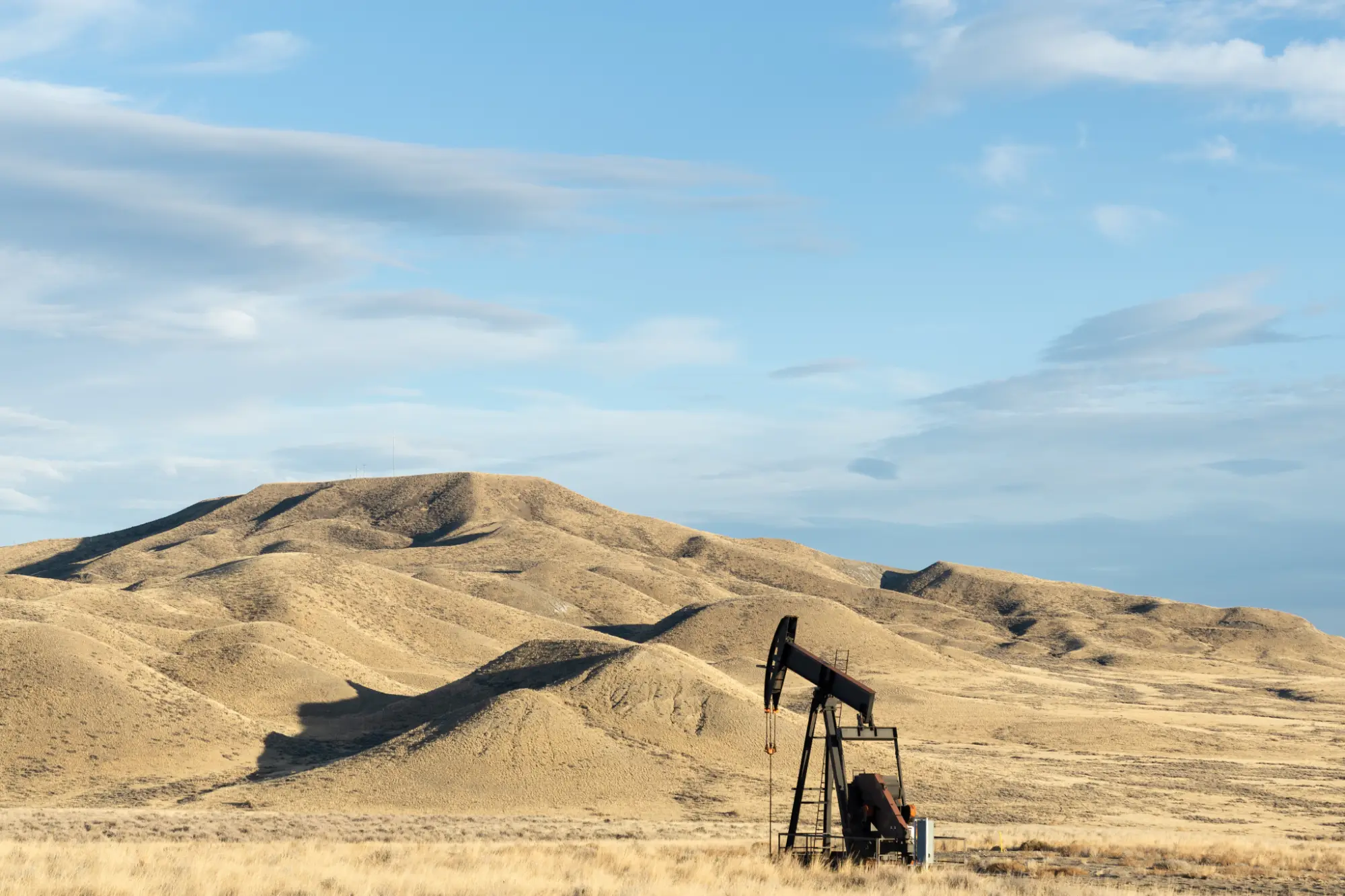 Oil pump jack operating in a dry, hilly landscape under a partly cloudy blue sky.