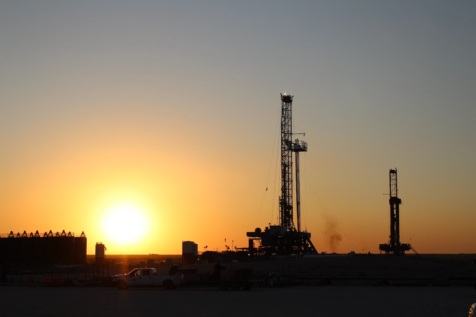 Oil drilling rigs silhouetted against a setting sun with a clear sky in the background.