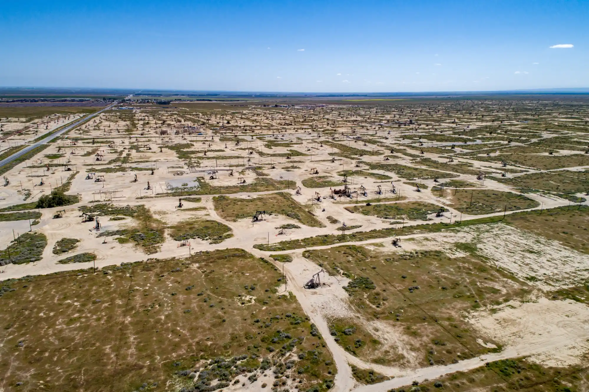 Aerial view of a vast oil field with numerous pumpjacks spread across a dry, flat landscape under a clear blue sky.