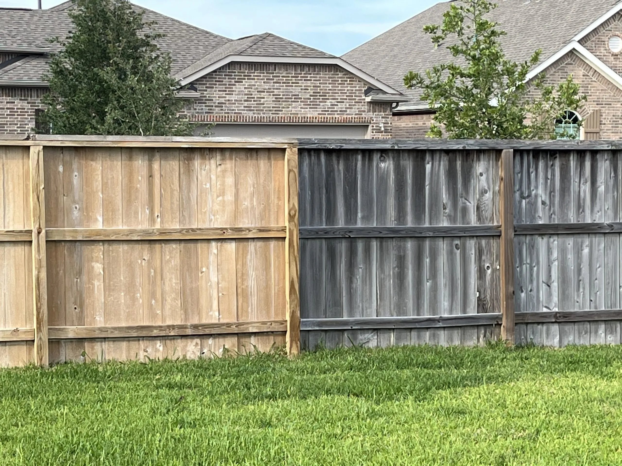 Two sections of wooden fence side by side, one new and light brown, the other old and weathered gray, with green grass in front and houses and trees in the background.