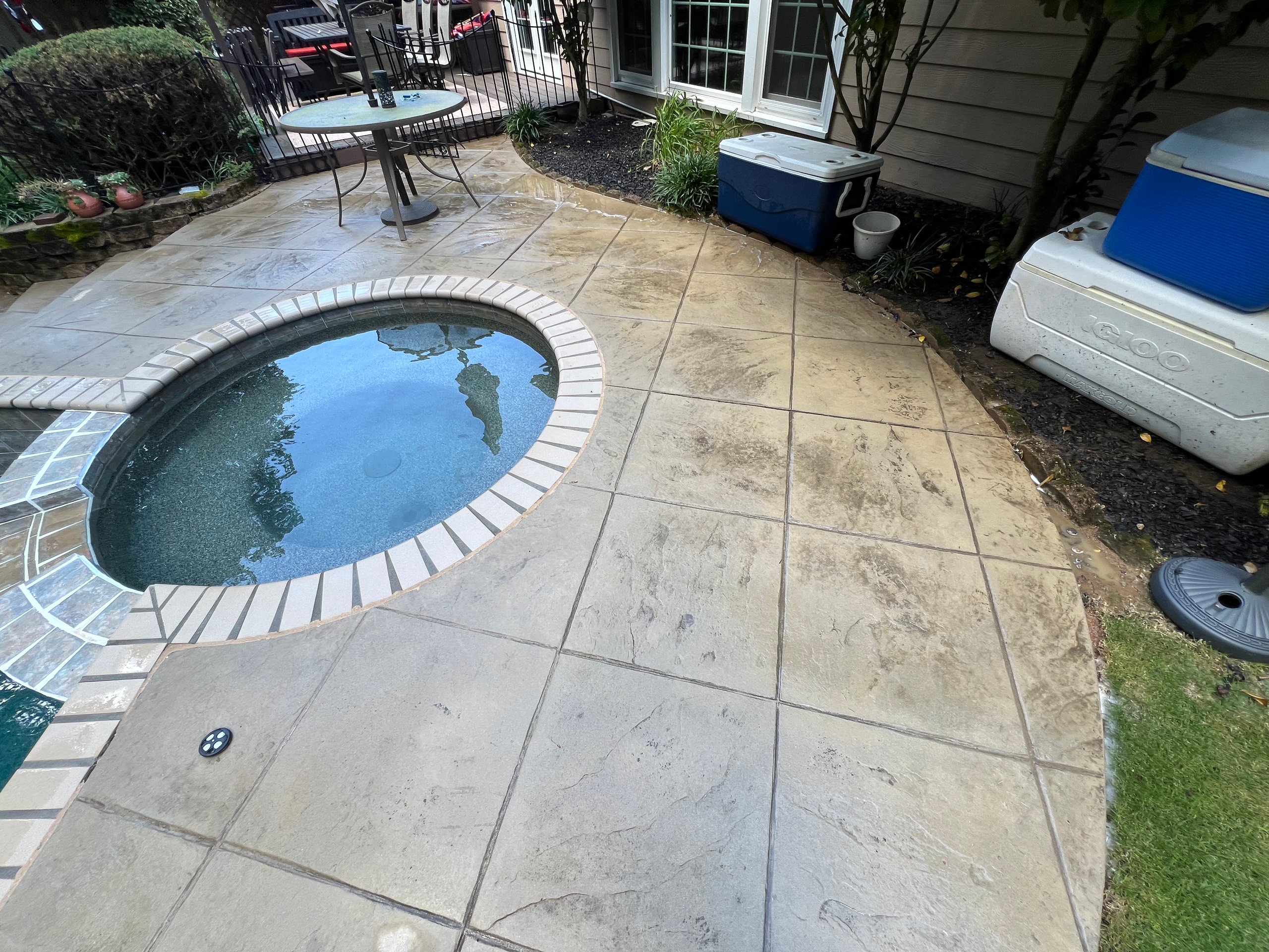Outdoor stone patio with a round hot tub, two coolers, and a glass-top table with metal chairs in a garden setting.