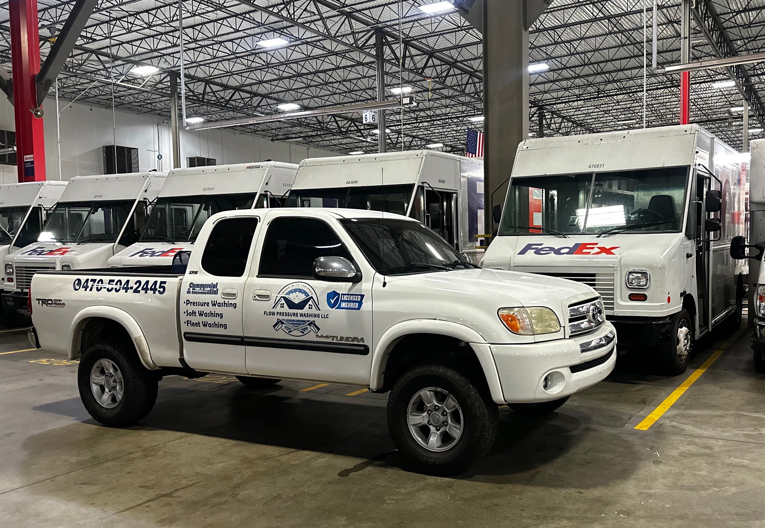 White Toyota Tundra pickup truck with Flow Pressure Washing LLC signage parked indoors in front of FedEx delivery trucks.