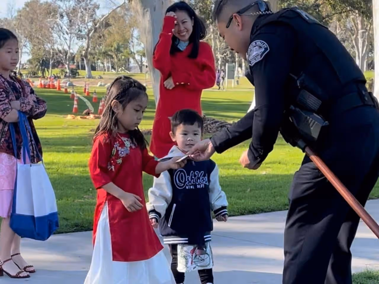 A police officer bending to hand an item to a young girl in a red dress, with a small boy and other children and a woman smiling in the park background.