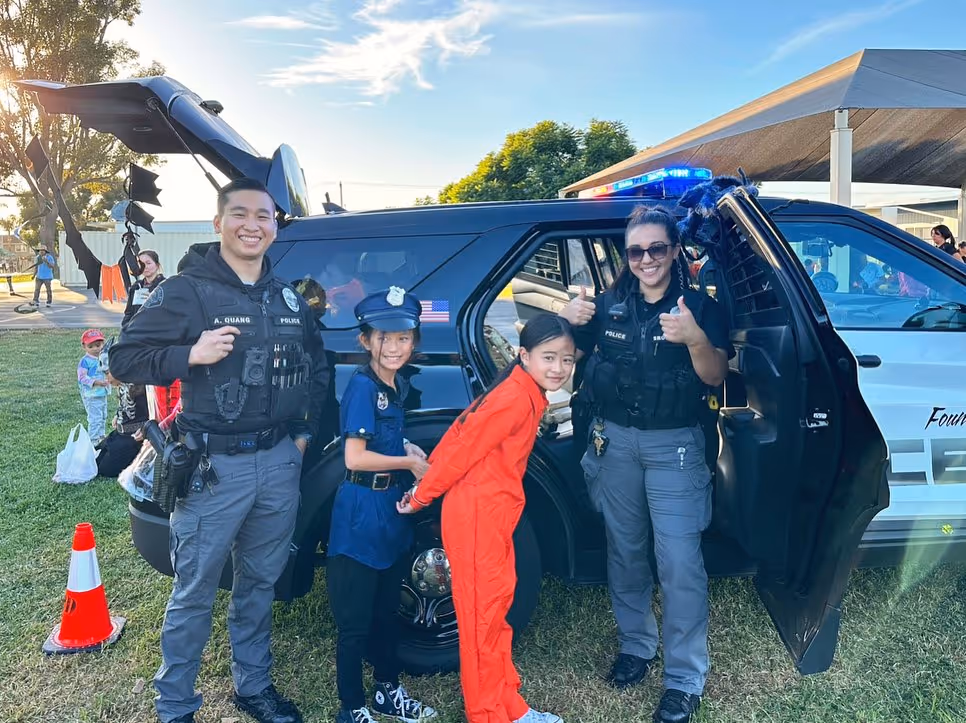 Two police officers smiling by a police SUV pose with two children dressed as a police officer and a prisoner during an outdoor community event.