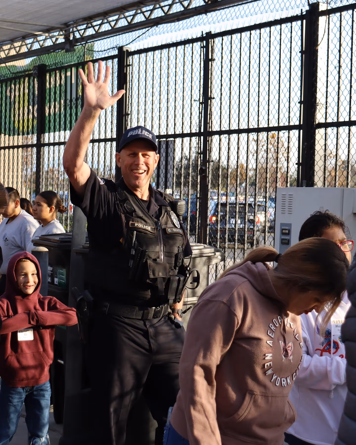 Smiling police officer in uniform waving with people standing nearby behind a chain-link fence.