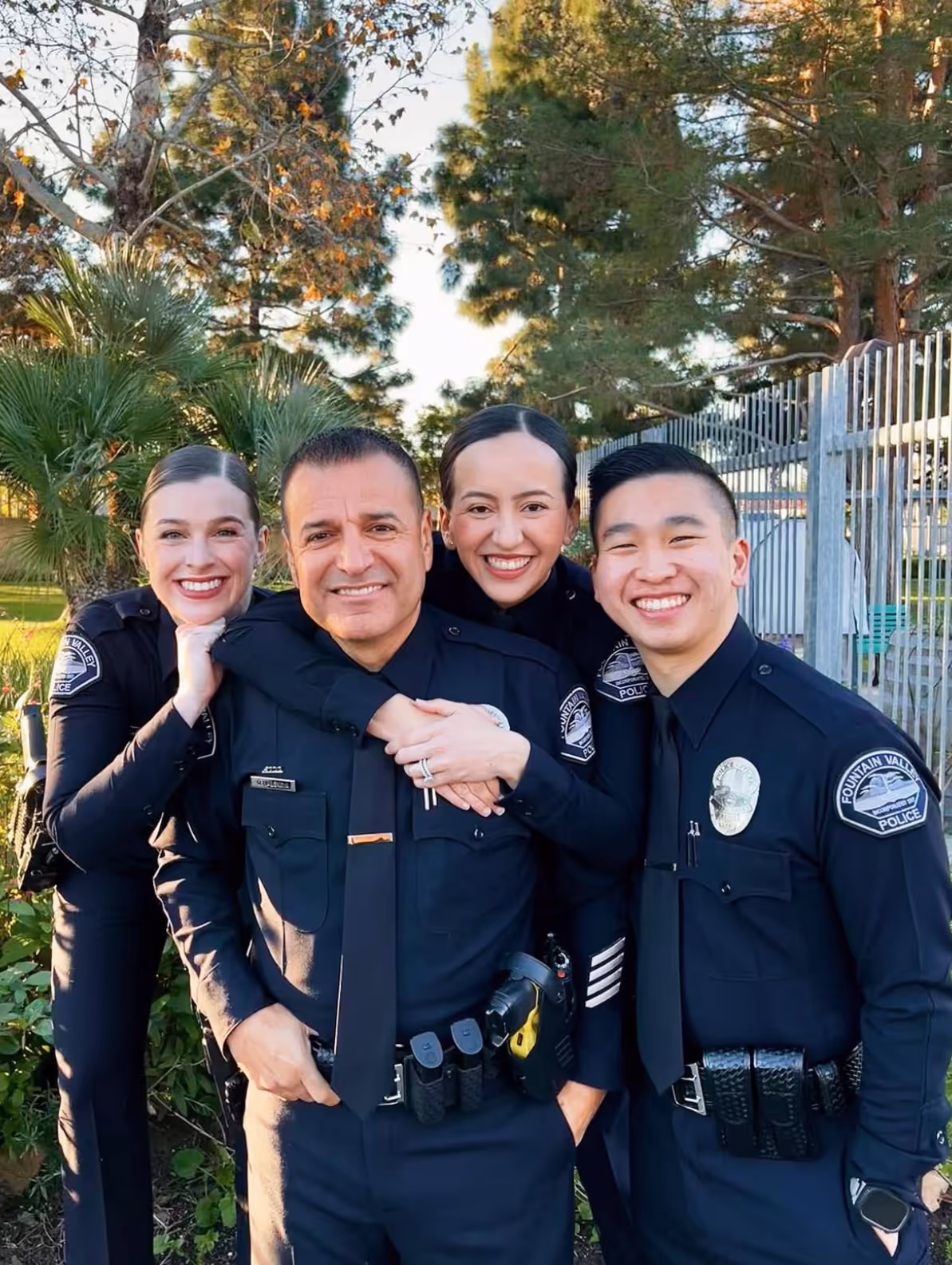 Four smiling police officers in uniform posing outdoors with trees and a fence in the background.