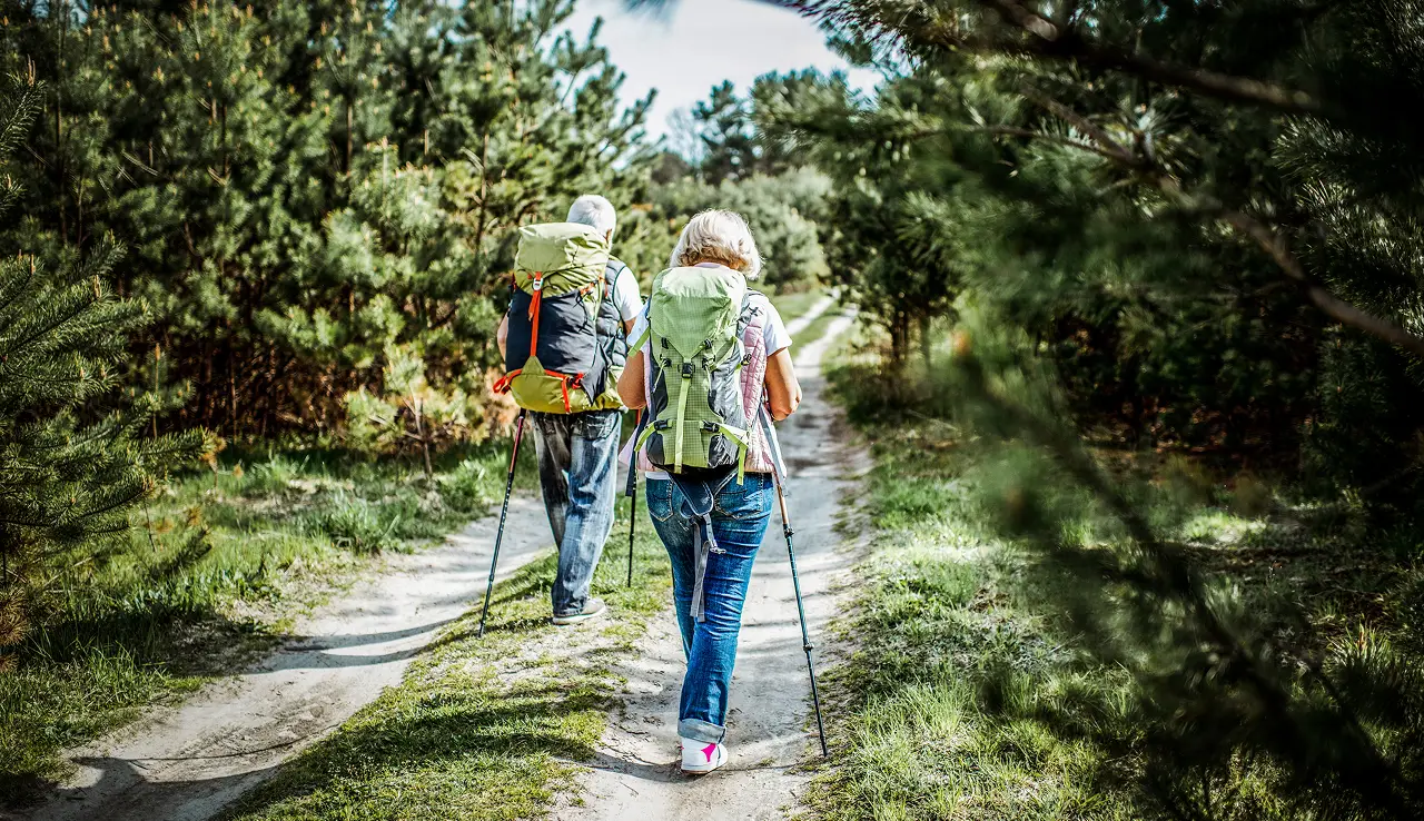Two retirees with backpacks hiking on a dirt trail through a forested area.