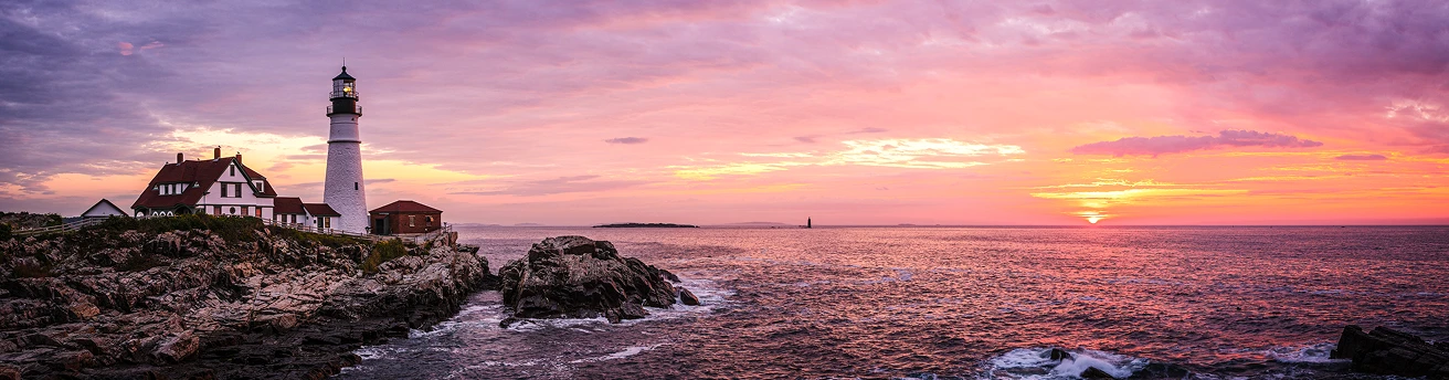 White lighthouse and buildings on rocky coast during purple and orange sunset over the ocean.