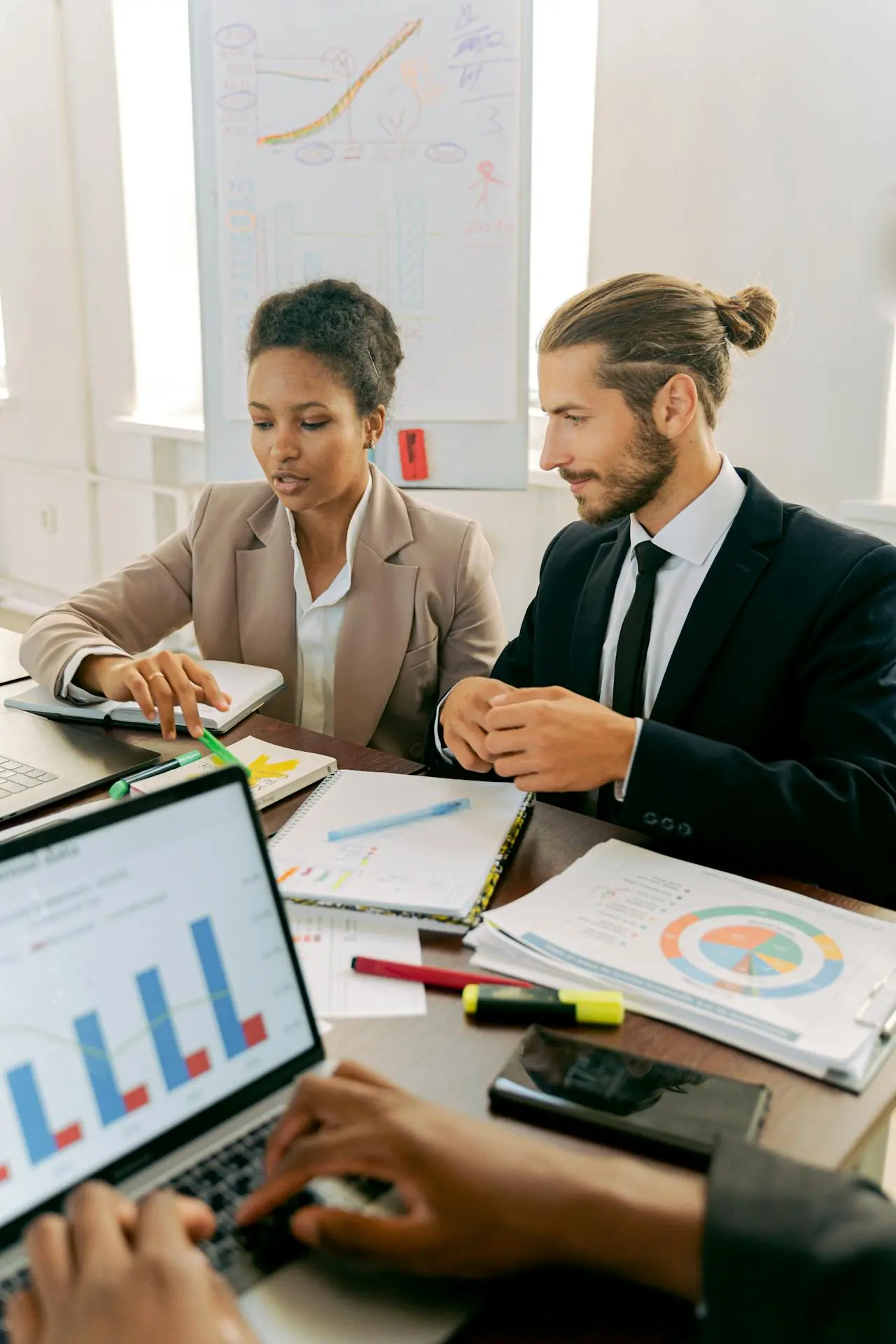 Two business professionals discussing documents at a table during a meeting with charts and a laptop.