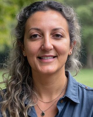 Smiling therapist woman with curly, graying hair wearing a dark blue shirt and a necklace with a green pendant standing outdoors.