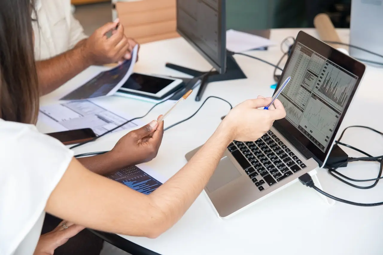 Two people collaborating at a desk with charts on a laptop and documents, pointing at the laptop screen.