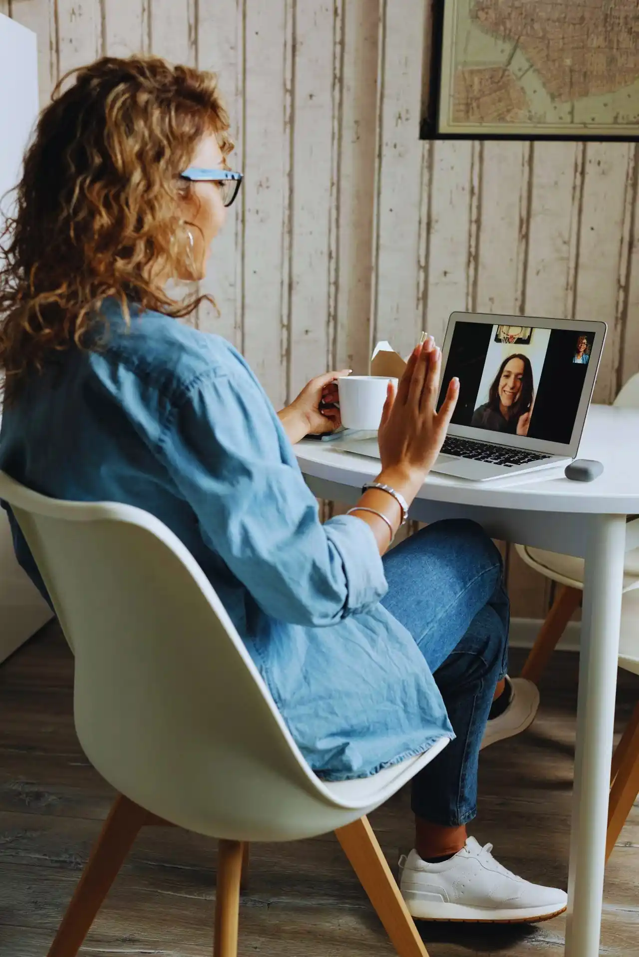 Woman sitting at a table holding a cup and waving during a laptop video call with another woman.