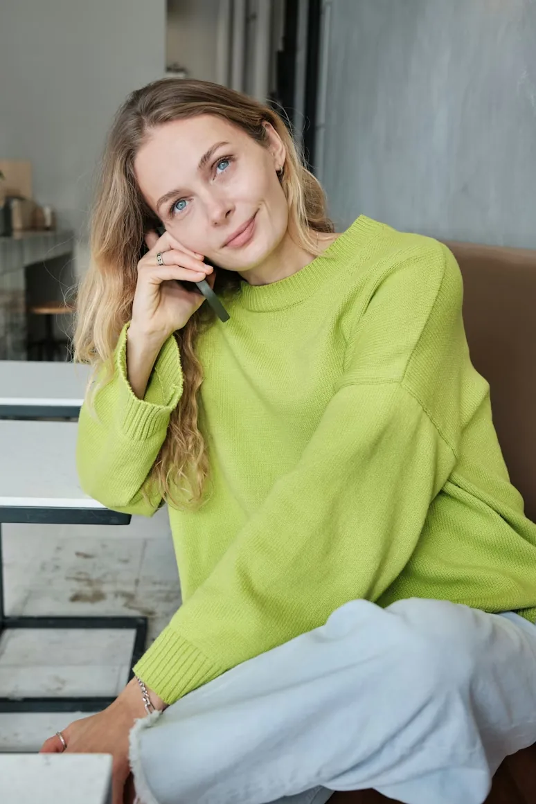 Young woman with blonde hair and blue eyes wearing a bright green sweater, sitting indoors and holding a phone to her ear.