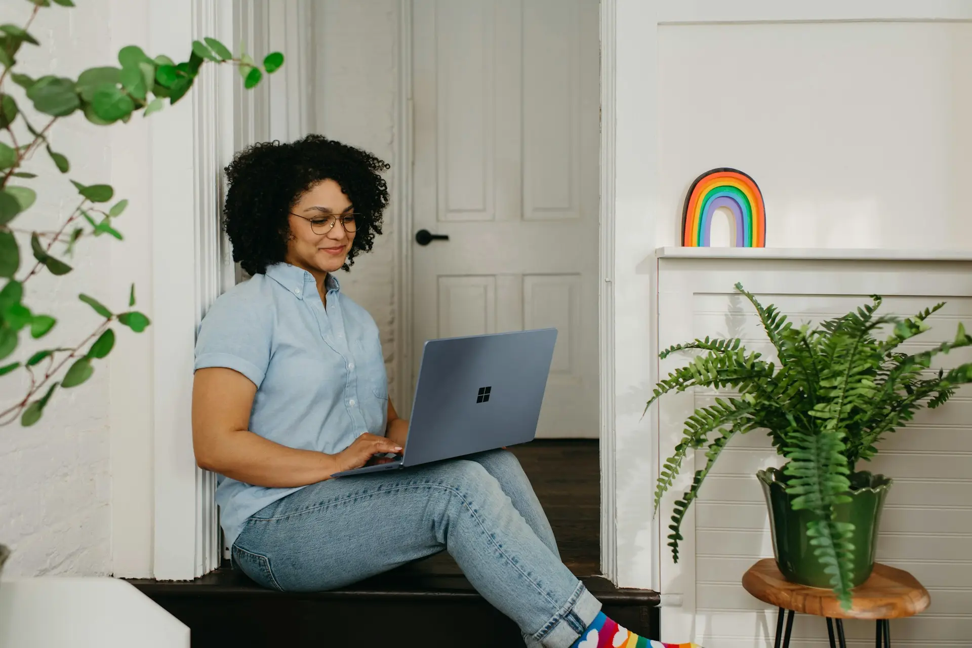 Smiling woman with curly hair sitting against a doorway using a laptop, wearing rainbow socks.
