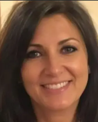 Smiling therapist woman with curly, graying hair wearing a dark blue shirt and a necklace with a green pendant standing outdoors.
