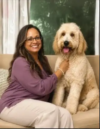 Woman smiling and sitting on a couch affectionately with a large curly-haired dog wearing a bandana.