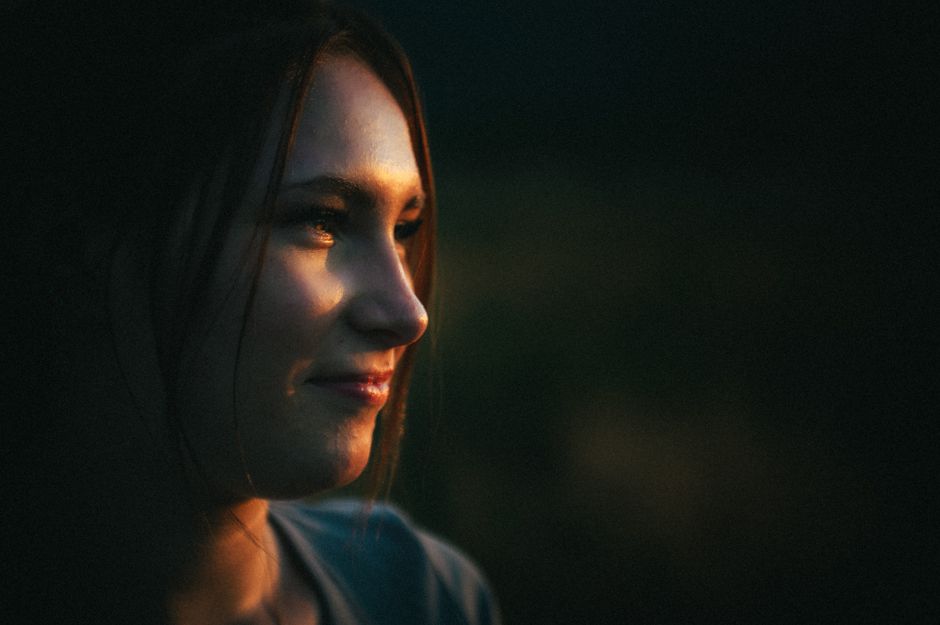 Portrait of a woman outdoors at golden hour, lit from the side and smiling faintly as she looks away from the camera against a dark blurred background