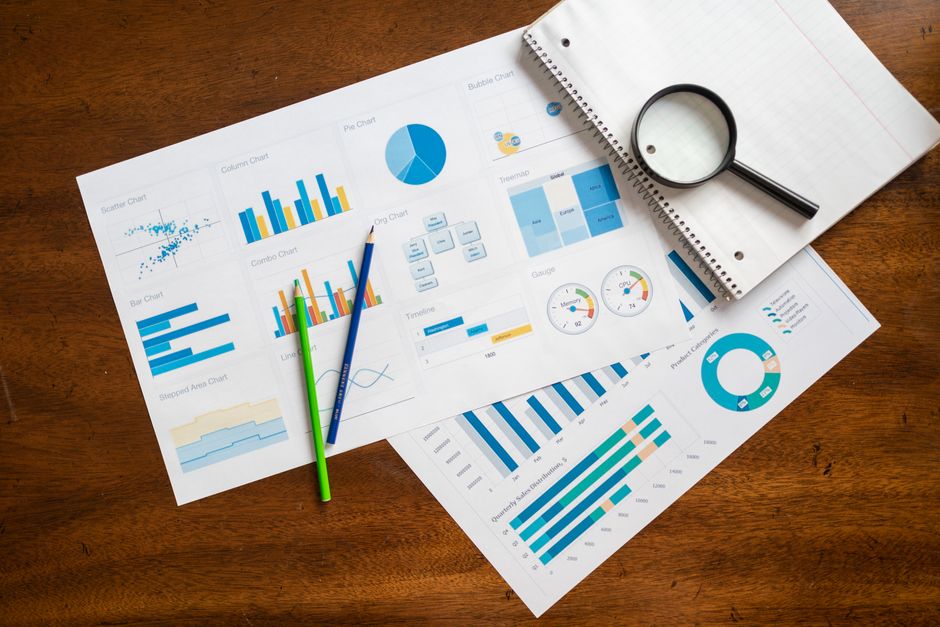 Printed marketing analytics reports spread on a wooden desk showing bar, pie, line and donut charts, with a magnifying glass, a notepad and two coloured pencils