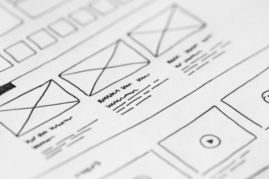 Hand-drawn paper wireframe of a web page layout, with sketched image placeholders marked by crossed boxes, navigation blocks and scribbled placeholder text lines