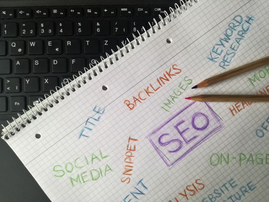 Spiral notebook resting on a laptop keyboard, covered in hand-written SEO terms including keyword research, backlinks, on-page, headlines and social media, circled around the word SEO