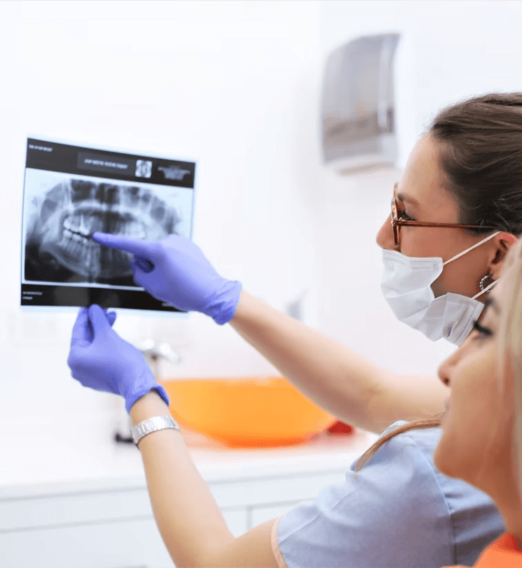 Dentista con guantes y barbijo señalando una radiografía dental mientras habla con una paciente en la clínica.