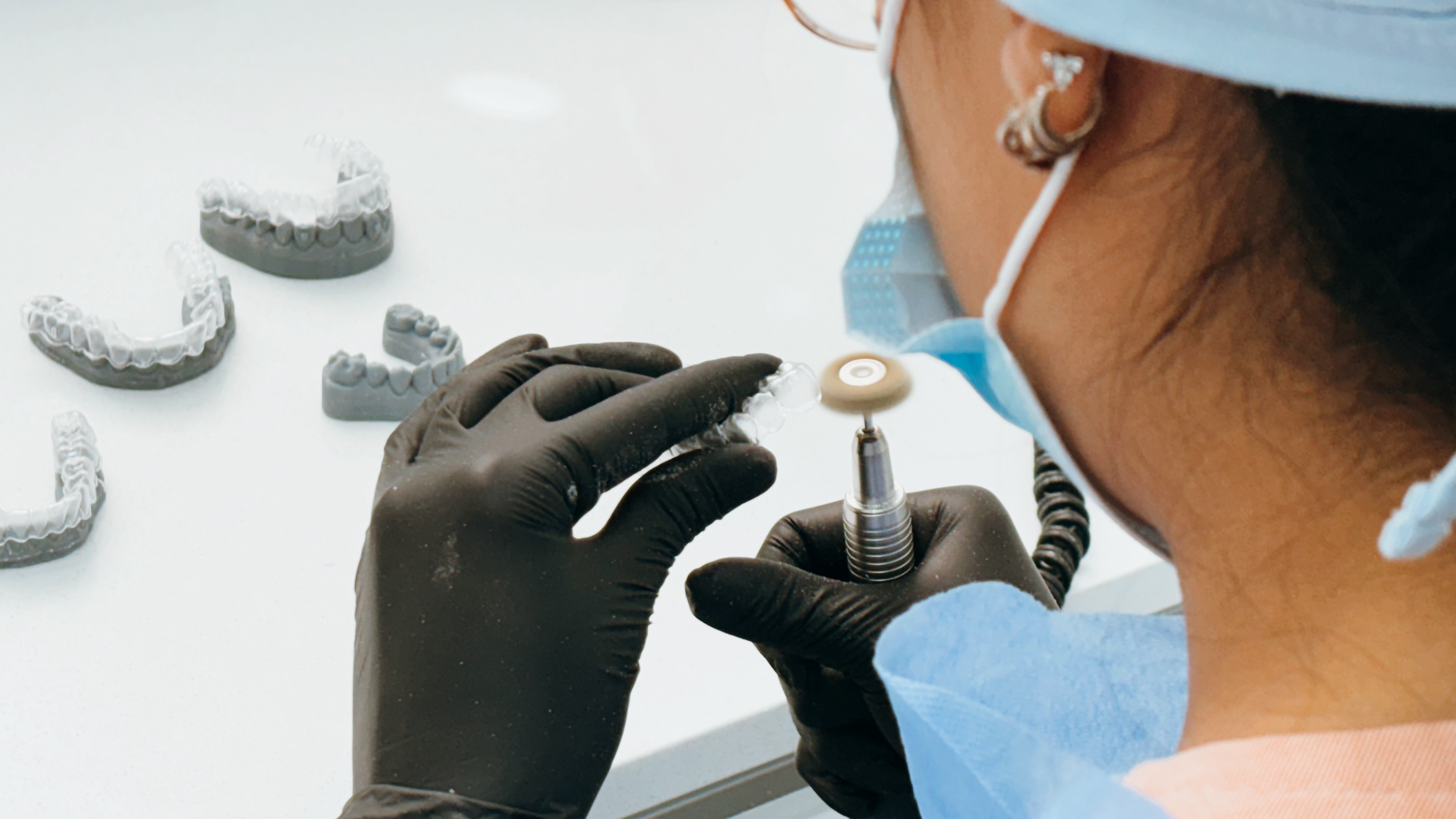 Profesional dental con guantes negros y mascarilla usando una pulidora para trabajar en una férula transparente sobre moldes dentales grises.