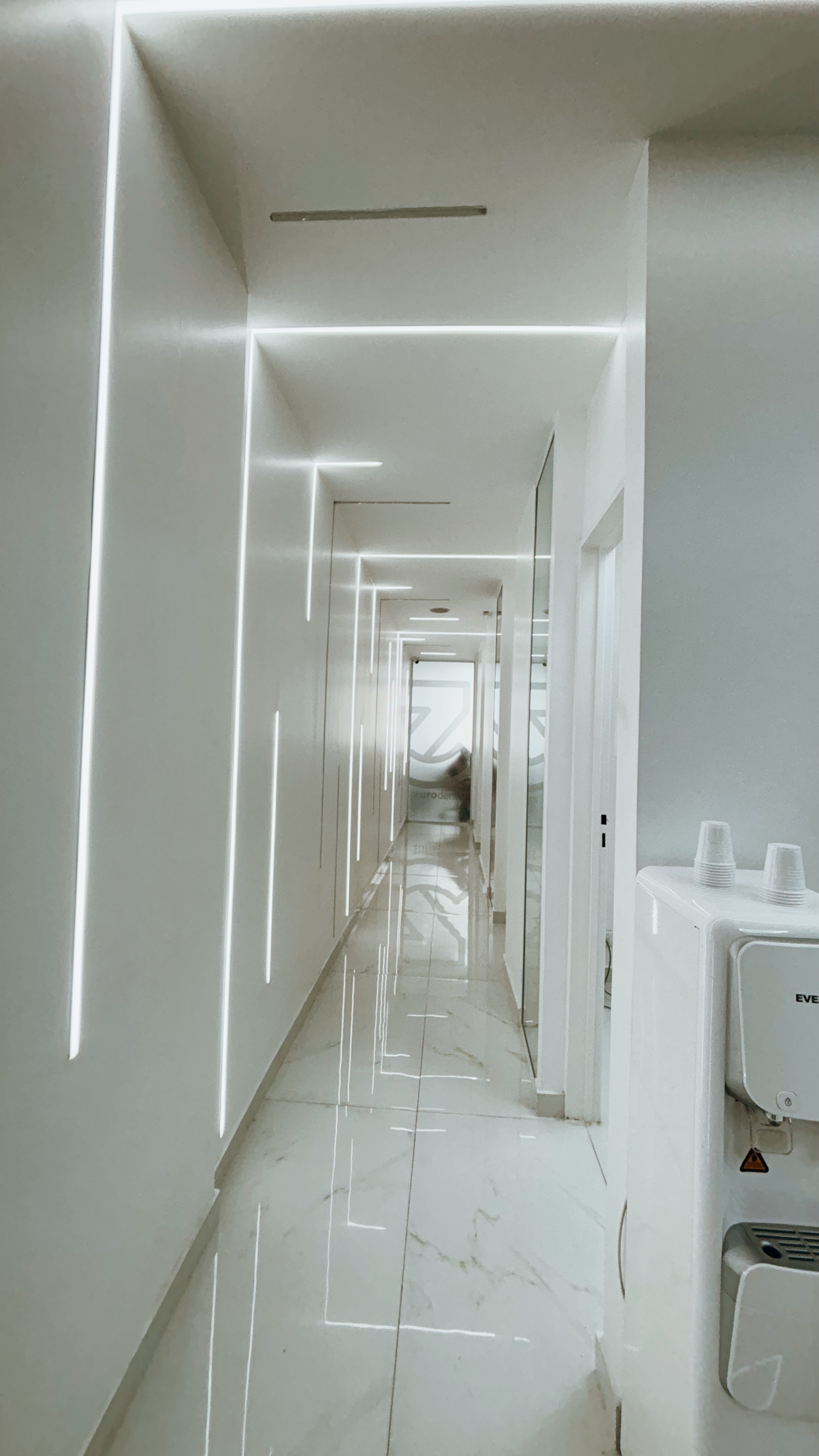 Pasillo largo y moderno con paredes blancas iluminadas por luces lineales y suelo de baldosas blancas brillantes.