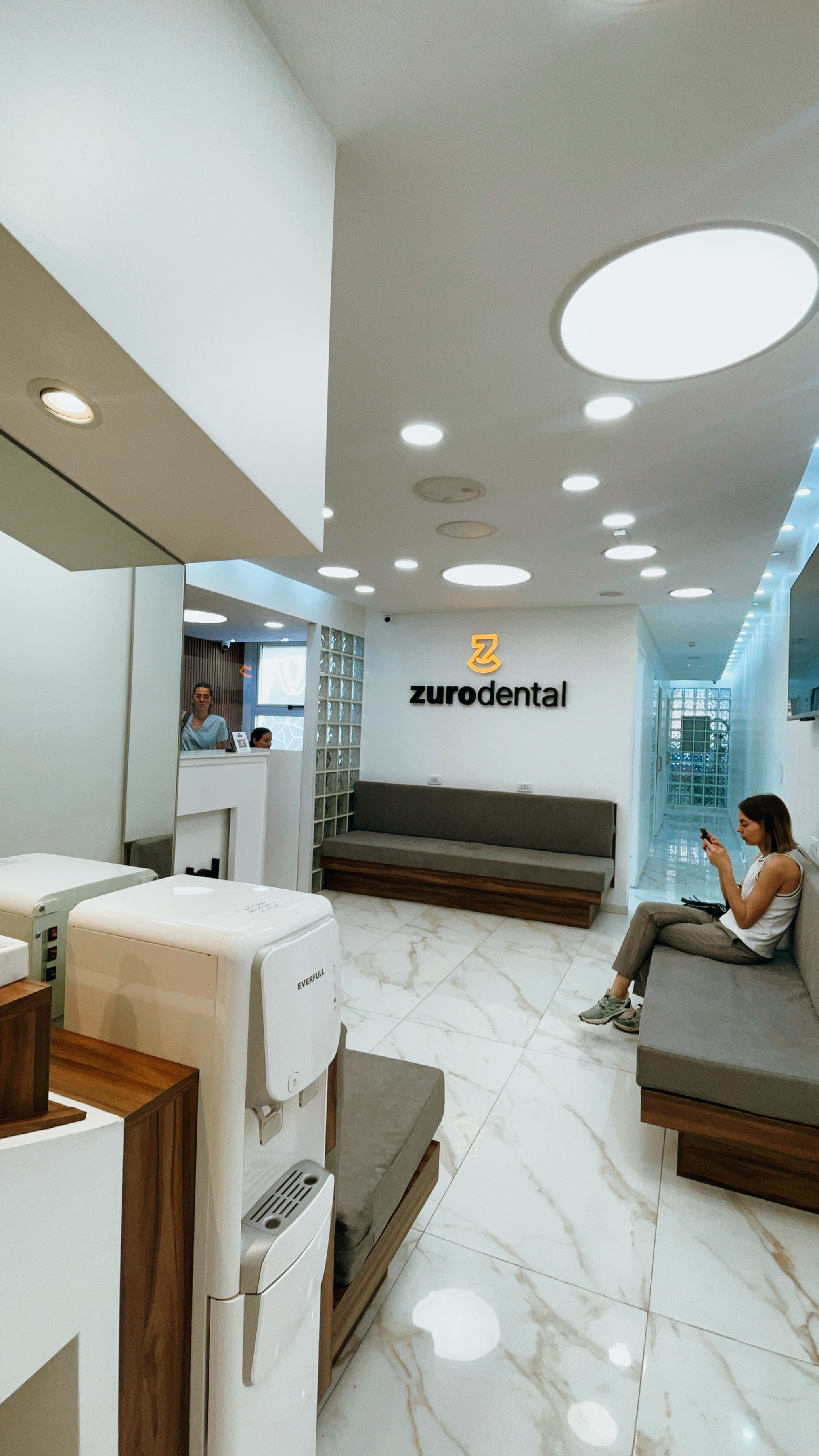 Sala de espera moderna de clínica dental ZuroDental con asientos grises, suelo blanco y mujer usando su celular.