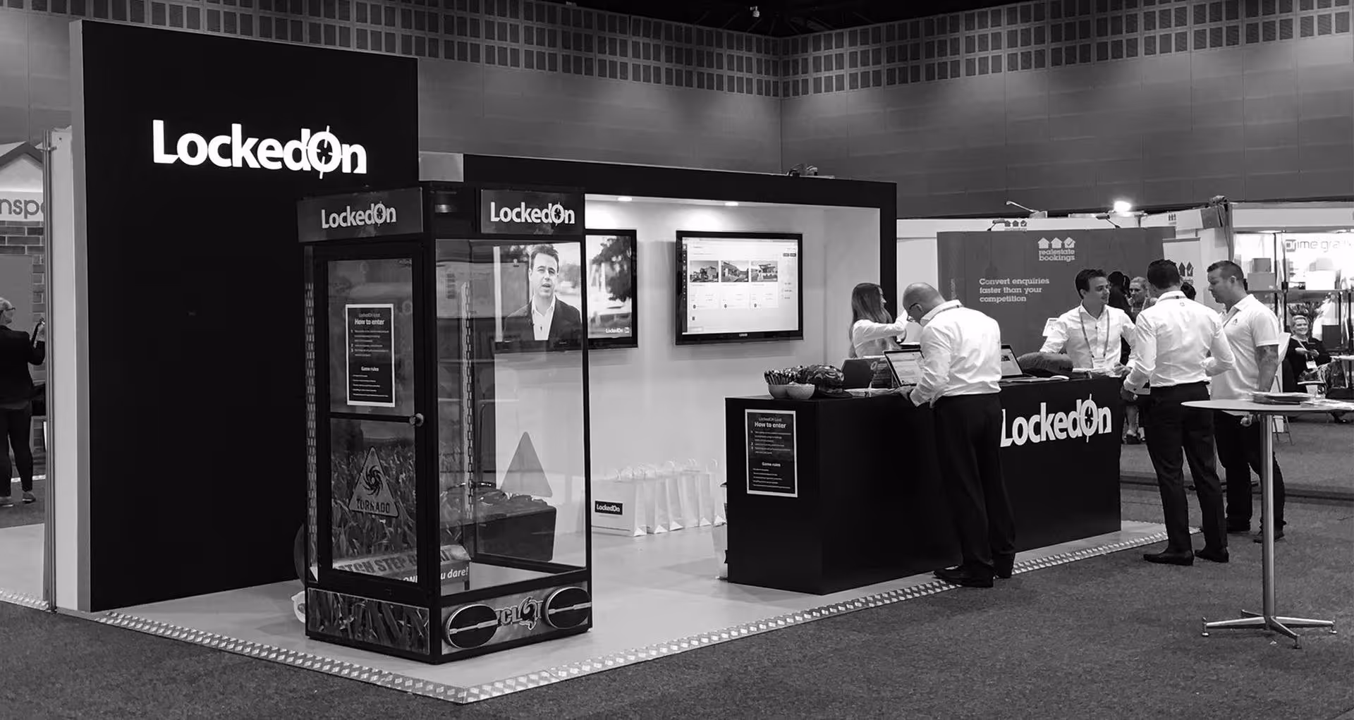 Trade show booth with LockedOn branding, featuring a glass display case, two wall-mounted screens, and staff interacting with visitors.