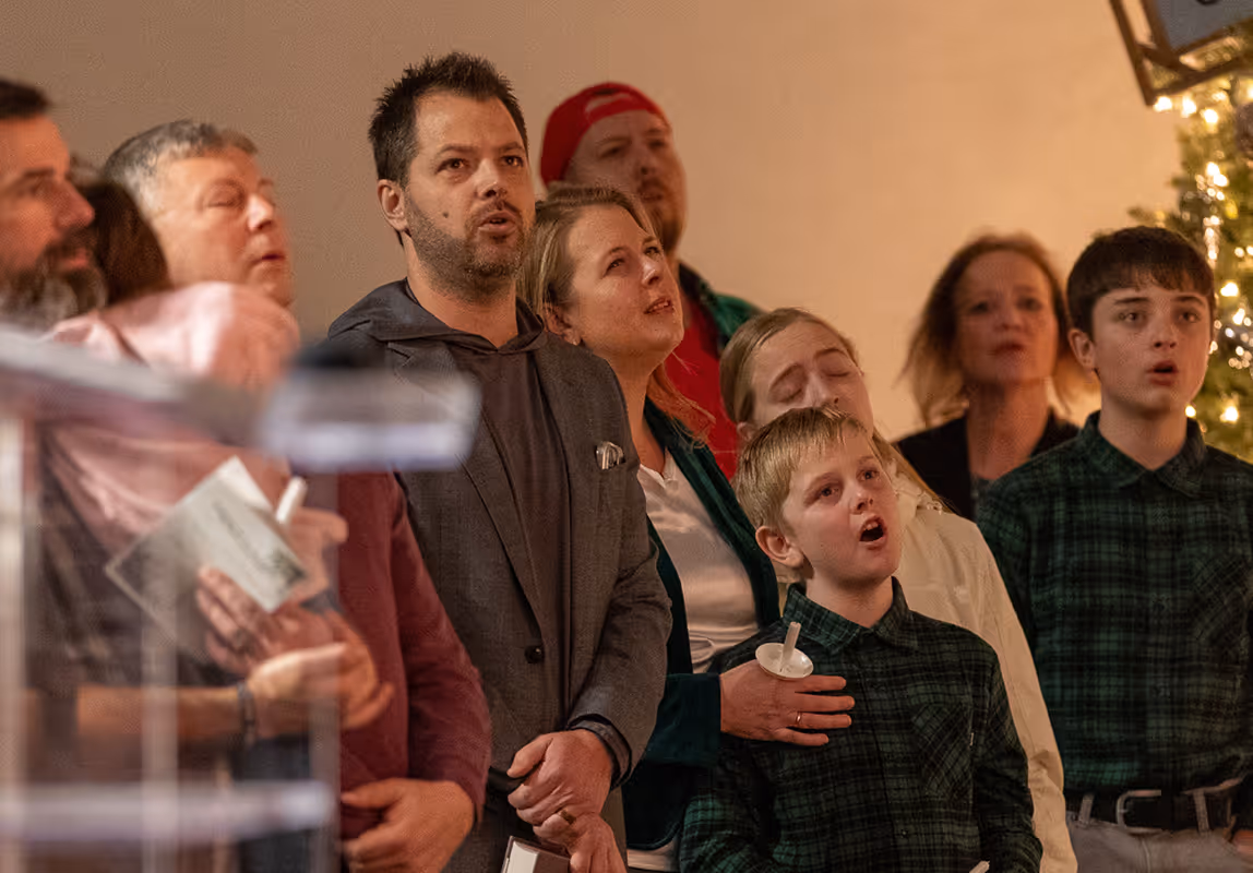 Group of people including adults and children singing together indoors near a decorated Christmas tree.