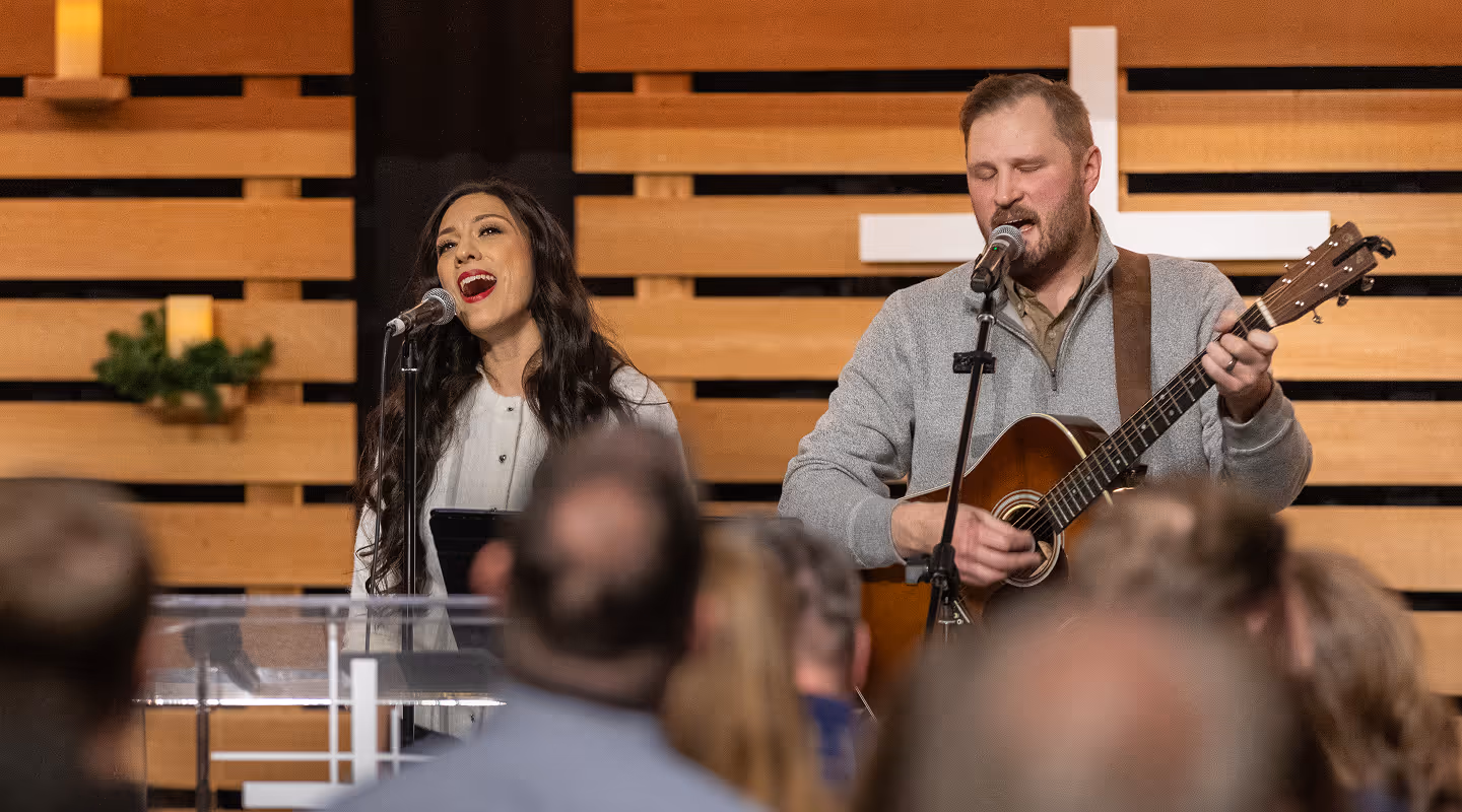 Man playing guitar and woman singing with microphones during a church service with a wooden cross in the background.