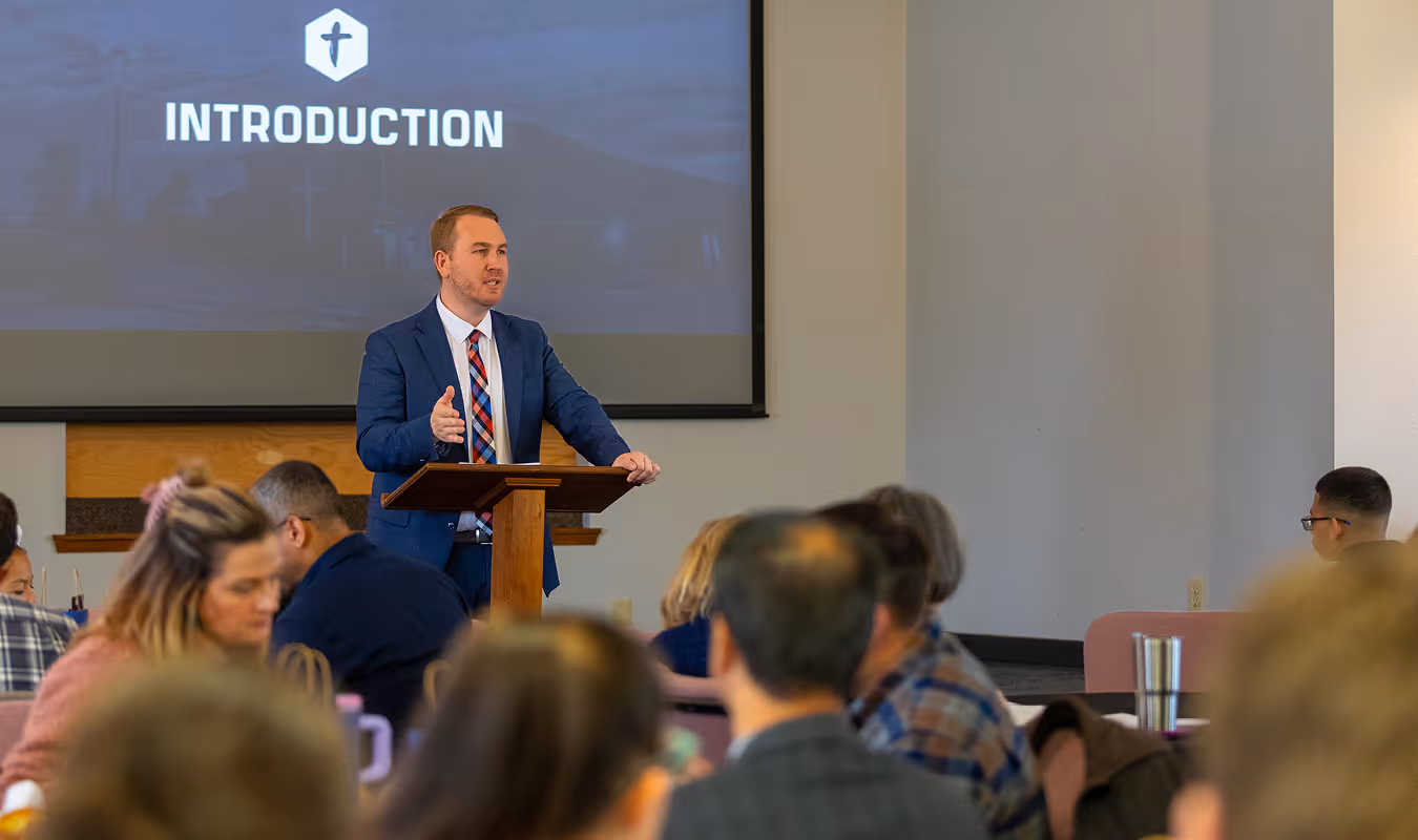 Man in a blue suit and patterned tie giving a presentation titled 'Introduction' to a seated audience in a conference room.