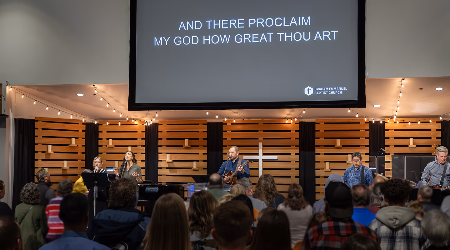 Congregation attending a church service with a worship band playing on stage in front of a wooden cross and a screen displaying the hymn lyrics 'AND THERE PROCLAIM MY GOD HOW GREAT THOU ART'.