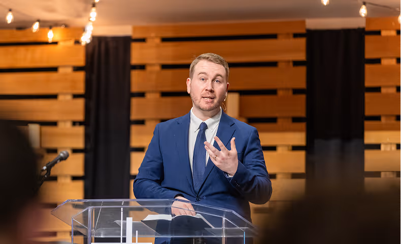 Man in blue suit speaking at a clear podium with a microphone and wood panel background.