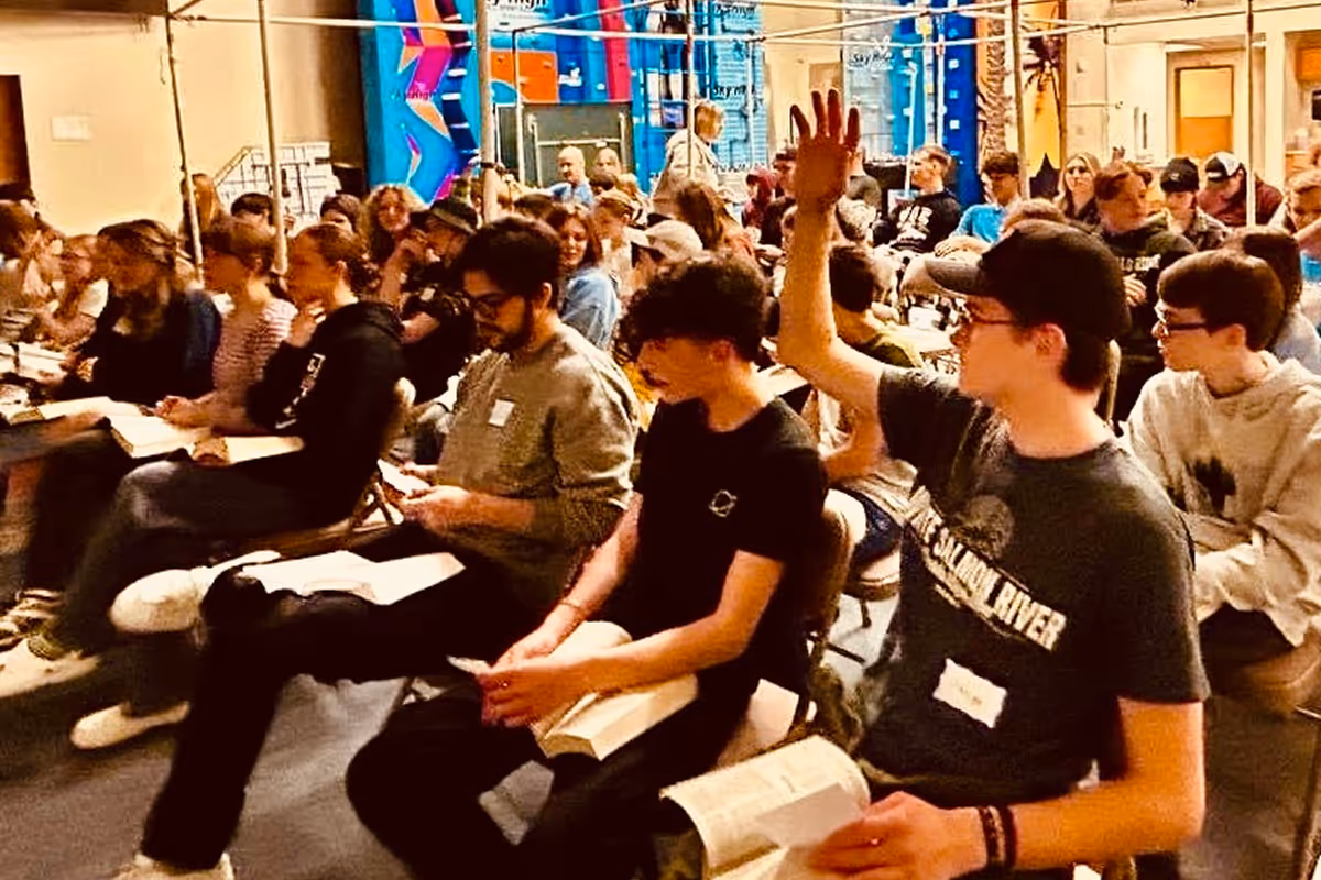Group of young people seated indoors, some reading books, one person raising their hand in a lively discussion setting.