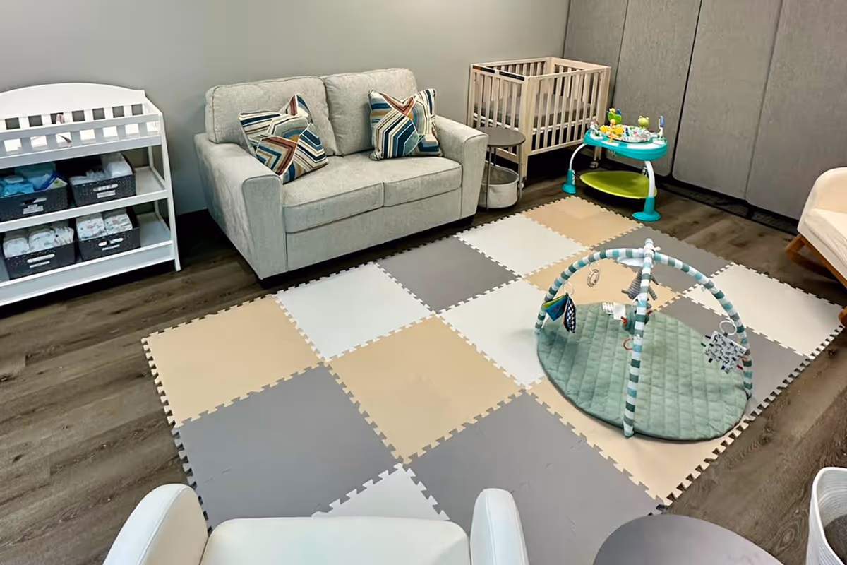 Modern nursery room with a gray couch, wooden crib, changing table with diapers, play mat with toy arch, and baby activity center.