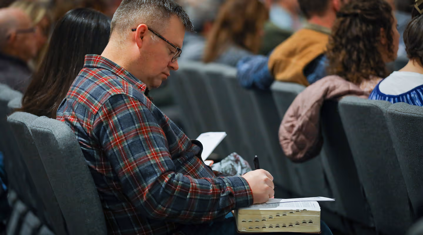 Man wearing glasses and a plaid shirt writing notes on paper placed on a thick book in a seated audience.