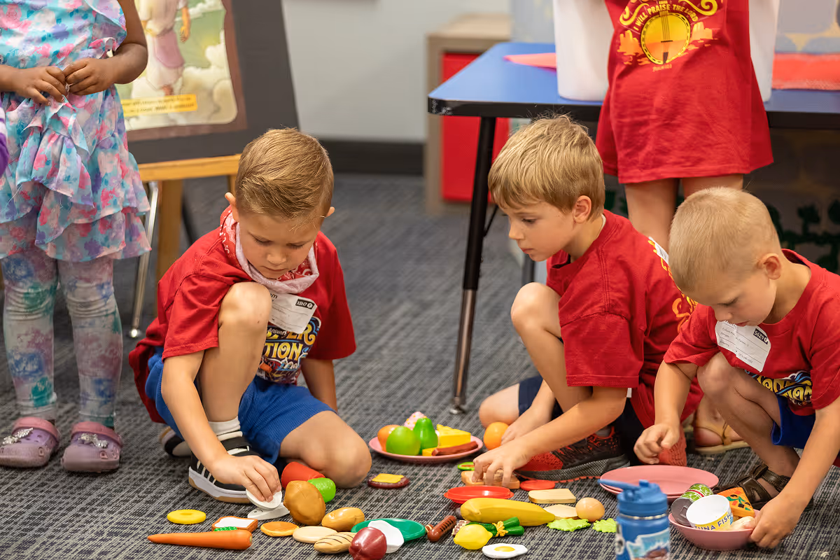 Three young boys in red shirts sitting on a carpeted floor playing with toy food items.